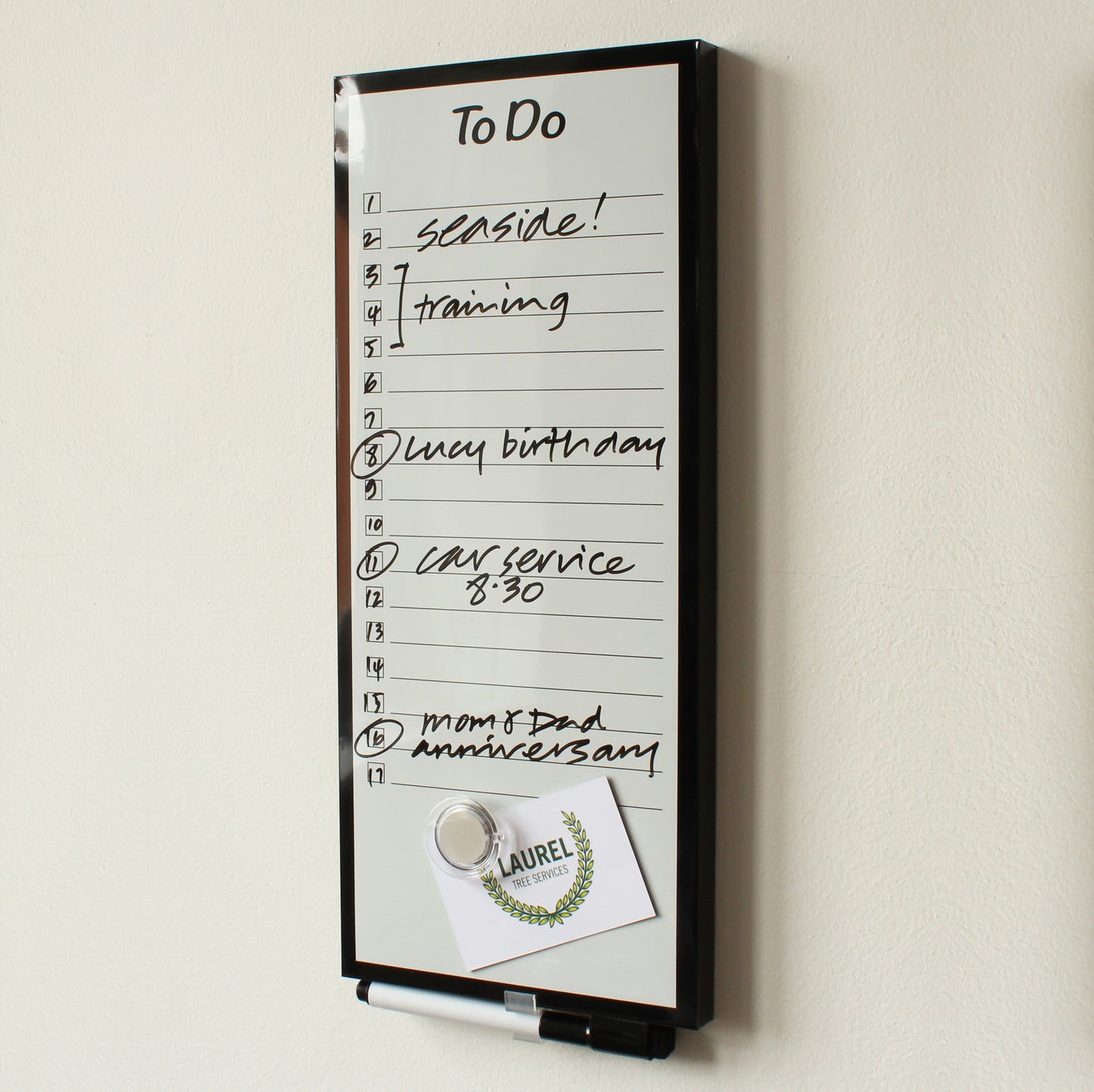 A frameless dry 'To Do' planner board (15 x 35 cm) mounted on a wall with tasks handwritten such as 'seaside!', 'training', 'Lucy birthday', 'car service 8:30', and 'mom & dad anniversary'. A promotional flyer from 'Laurel Leaf Services' with a laurel wreath design is attached to the board using a round, transparent magnet and a black dry erase marker with eraser in the lid is clipped into the pen holder at the top of the board.