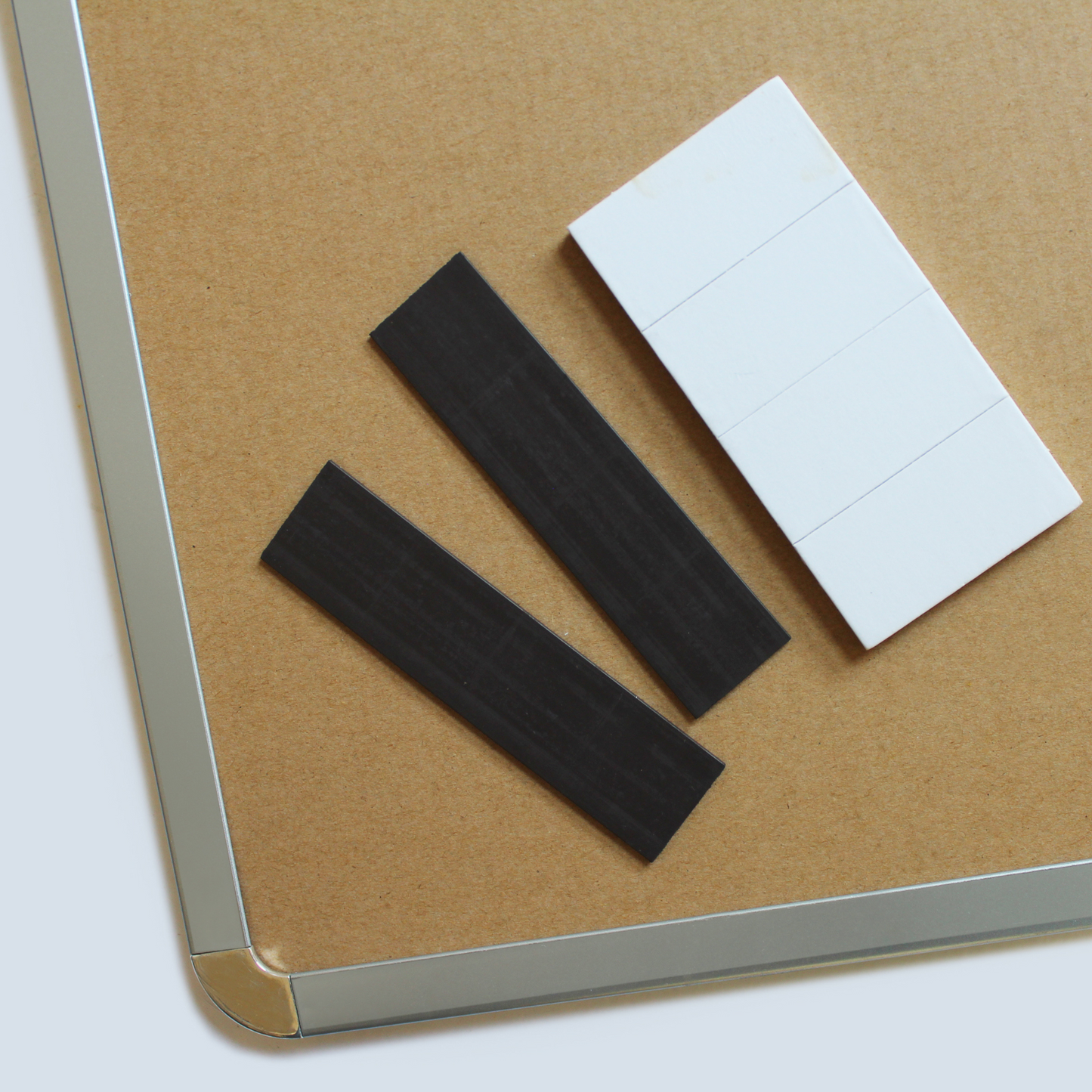 Close-up of the rear of a dry erase board with chrome corners, sized 28x43cm, showcasing two strips of black magnetic tape and a stack of white sticky pads, showing 2 ways of mounting the board, either using the included sticky pads, or magnets to be affixed to a magnetic surface, such as a fridge.