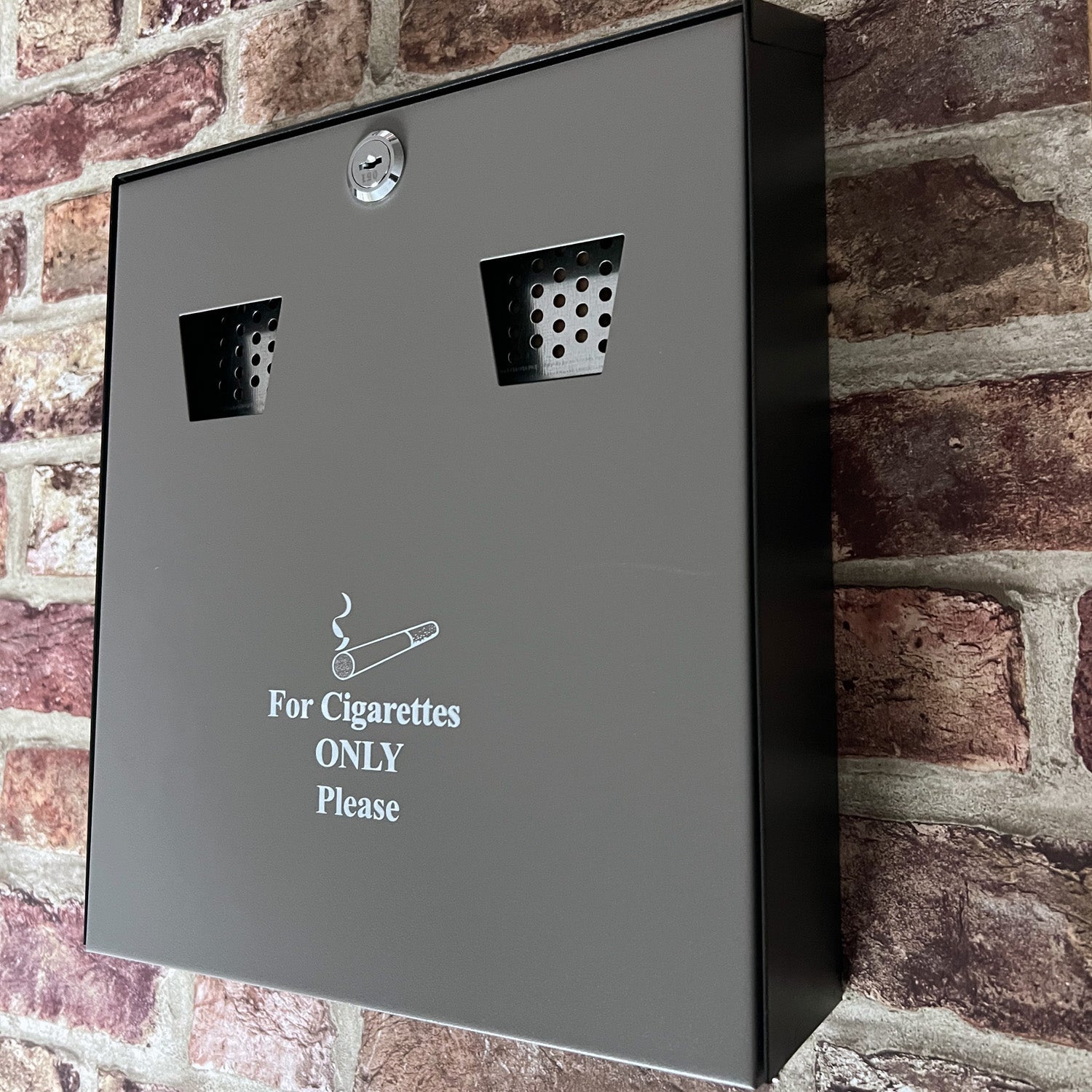 Wall-mounted ash bin with a 3.1L capacity, attached to a brick wall. The bin is dark grey with a lock at the top and features two stub plates. It is labeled 'For Cigarettes ONLY Please' to designate its purpose.