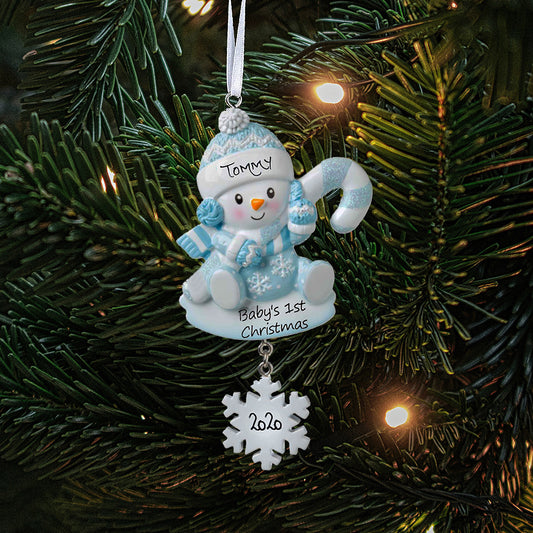 Personalized baby Christmas decoration featuring a blue snowbaby holding a blue striped candy cane while wearing a blue bobble hat, with name 'Tommy' written on his hat, and '2020' written from a hanging snowflake, showing the personalisation aspect of the decoration, the decoration also has 'Baby's 1st Christmas' printed on the base. The ornament is shown against a festive tree background.