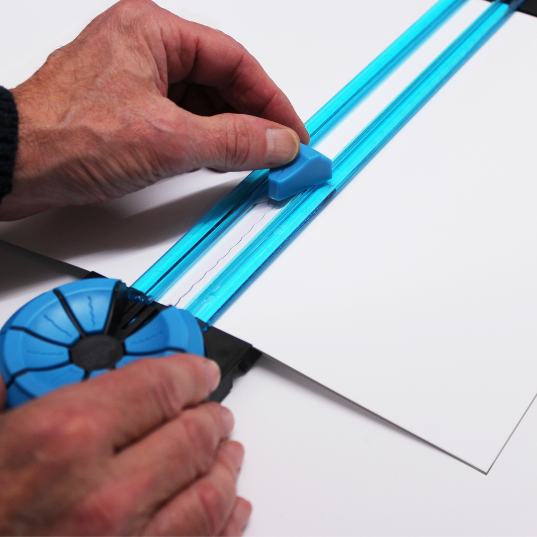 Hands operating the A4 Dial-A-Blade Creative Trimmer on a white sheet of paper, showcasing the trimmer's blue dial and cutting head in action, illustrating the ease of changing between its functions for precise paper crafting.