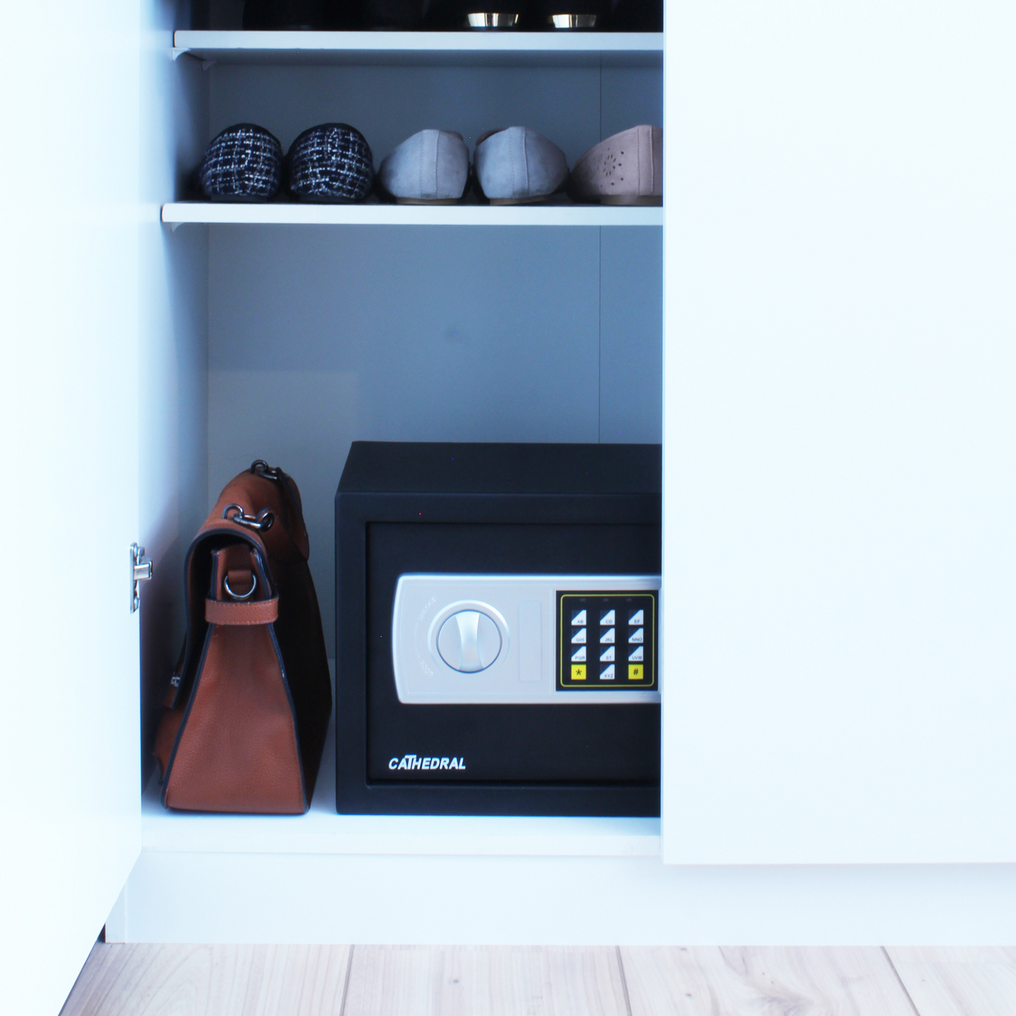A Cathedral Products EA25 16 Litre Electronic Digital Safe with manual override is neatly tucked in a white cupboard beside a brown messenger bag, illustrating a practical and discreet home security solution.