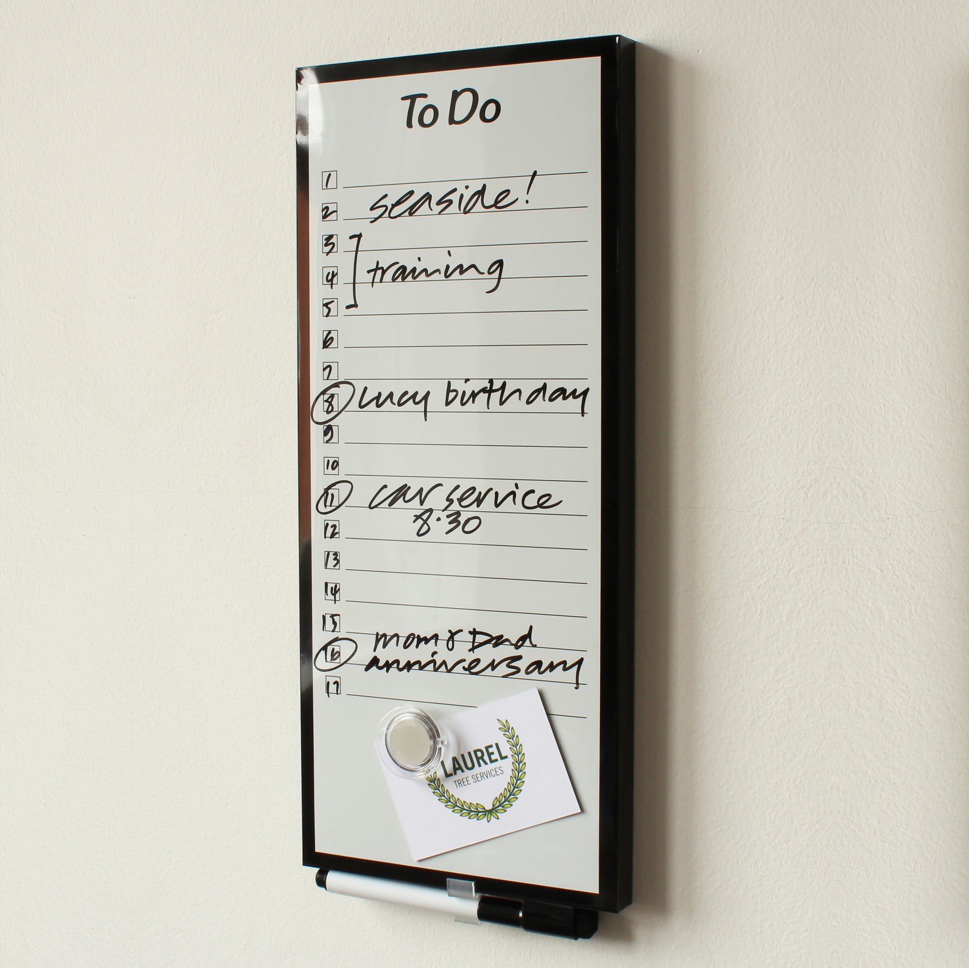 A frameless dry 'To Do' planner board (15 x 35 cm) mounted on a wall with tasks handwritten such as 'seaside!', 'training', 'Lucy birthday', 'car service 8:30', and 'mom & dad anniversary'. A promotional flyer from 'Laurel Leaf Services' with a laurel wreath design is attached to the board using a round, transparent magnet and a black dry erase marker with eraser in the lid is clipped into the pen holder at the top of the board.