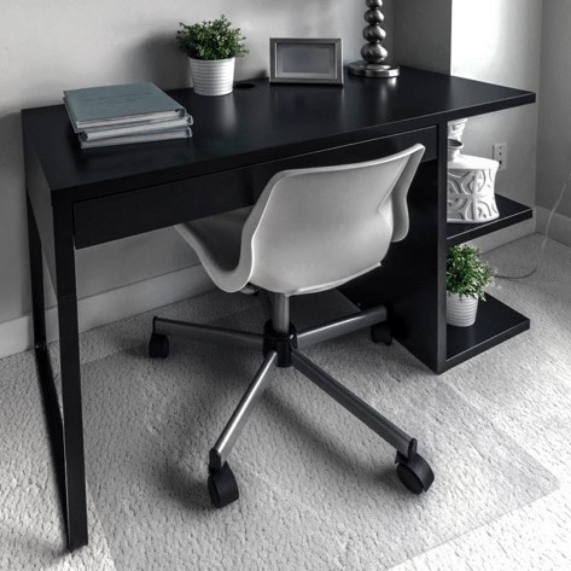 A contemporary home office setup with a black desk and a white modern office chair on a light carpeted floor, with a chair mat protecting the carpet from indents caused by the chair. The desk is adorned with neatly stacked books, a potted plant, and decorative items, creating a clean and stylish workspace.