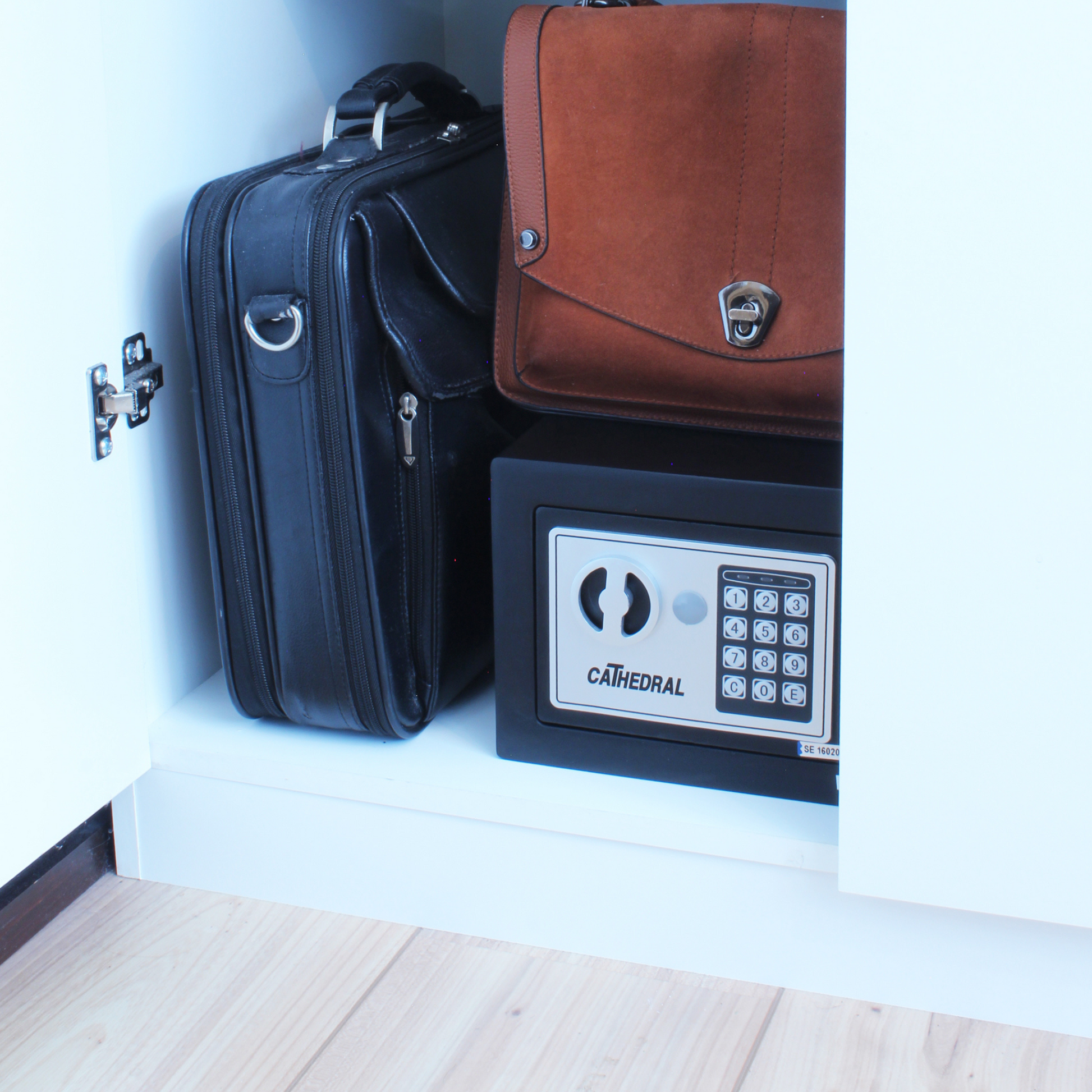 A Cathedral Products EA15 5 Litre Electronic Digital Safe with manual override is neatly tucked in a white cupboard beside a black briefcase and a brown messenger bag, illustrating a practical and discreet home security solution.