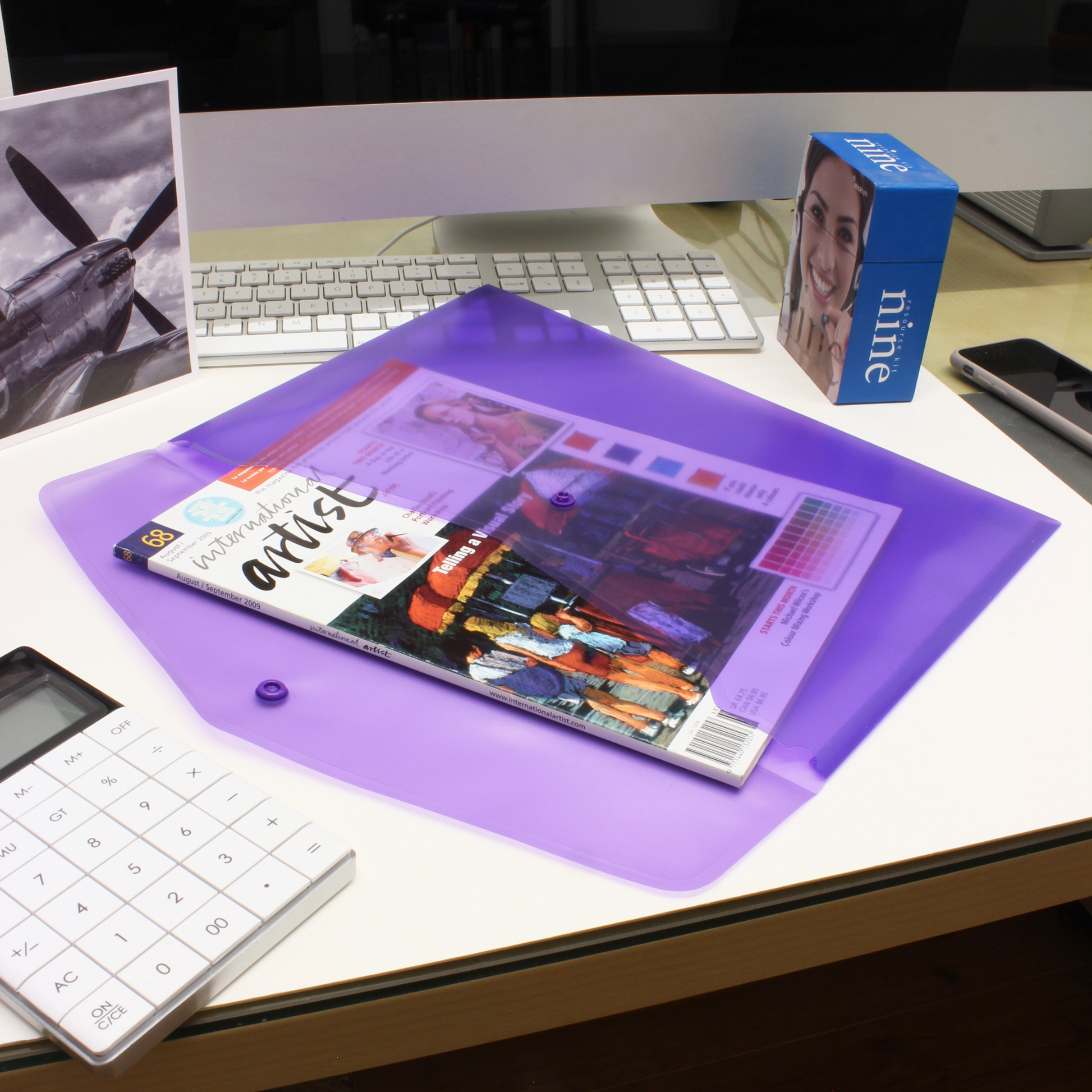 Purple A4 plastic stud wallet on a workspace, displaying a vivid magazine cover, next to a calculator with a black-and-white airplane photo and keyboard in the background, illustrating an organized desk with stationery essentials.