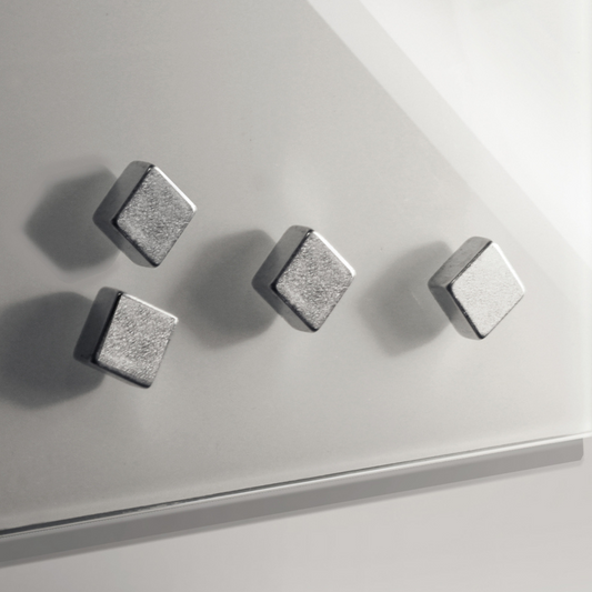 Four square-shaped, designer neodymium magnets with a sleek silver finish are arranged diagonally on a glass surface, casting soft shadows due to the lighting.