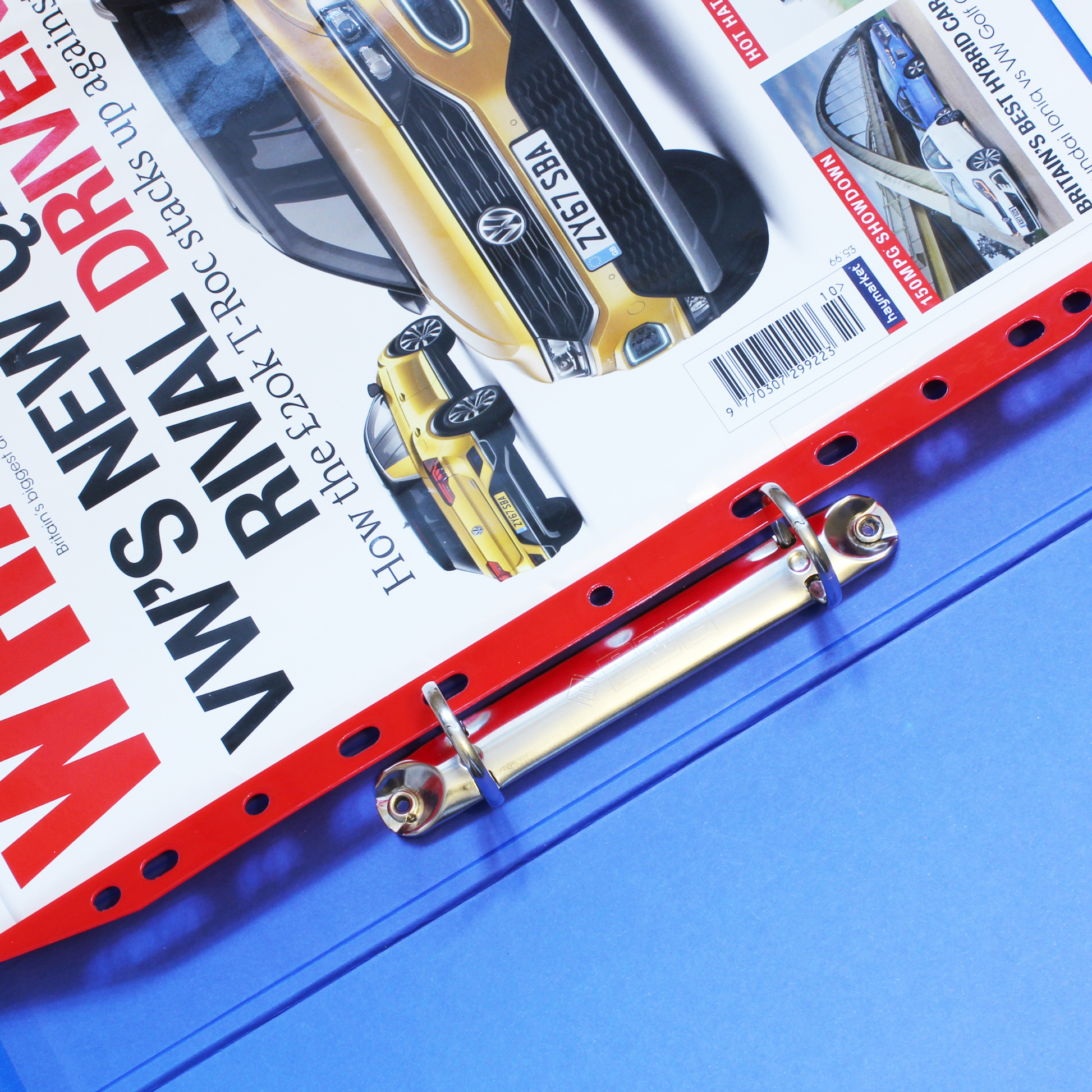 A close-up of a red Magi Clip multi-punched magazine storage clip, in situ in a ring binder, holding an issue of 'WHAT CAR?' magazine, showcasing a yellow sports car on the cover.