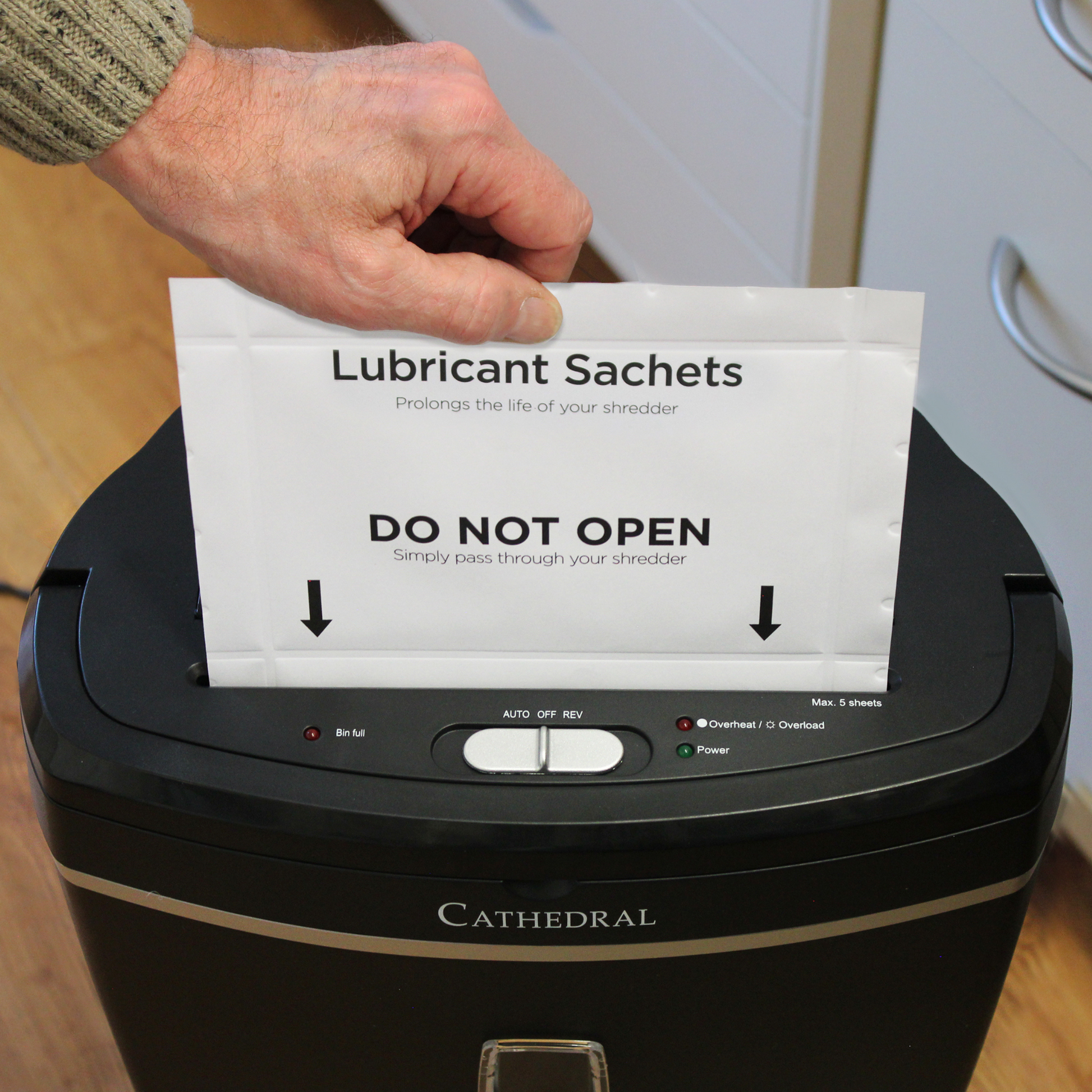 A person's hand is holding a lubricant sachet labeled 'DO NOT OPEN - Simply pass through your shredder' above a black paper shredder that reads 'Cathedral' on the front. The sachet is intended to prolong the life of the shredder.