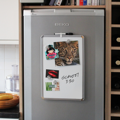 A functional 28x43cm dry erase board with chrome corners, attached to a BEKO fridge door, adorned with a colorful cartoon figure, an intense leopard photo, a red lips card, and a business card for 'Clinic 20', with a handwritten reminder 'School 3:30'. The image conveys that the dry erase board can be affixed to a fridge, using the included magnetic tape.