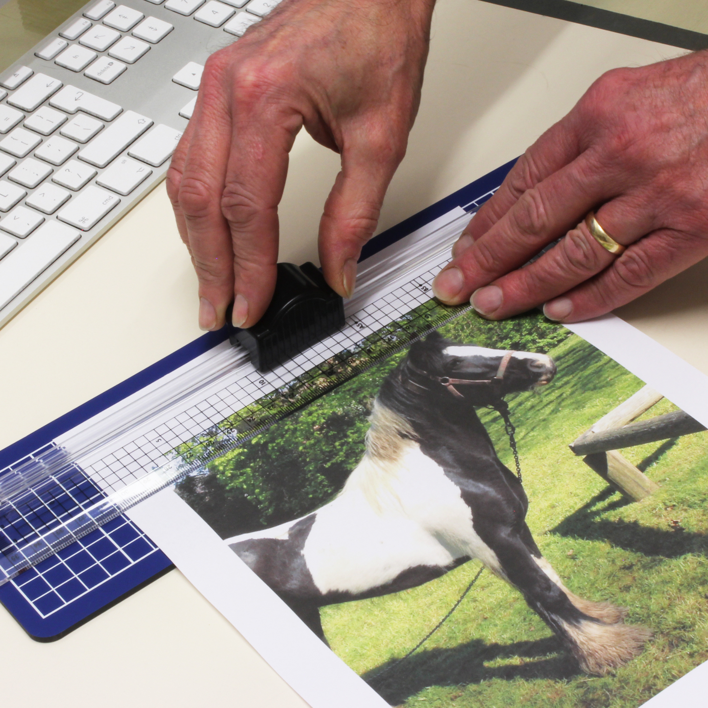 Hands using a transparent cutting ruler with a black grip to trim a photo of a horse on an A4 blue cutting mat, with a computer keyboard in the background.