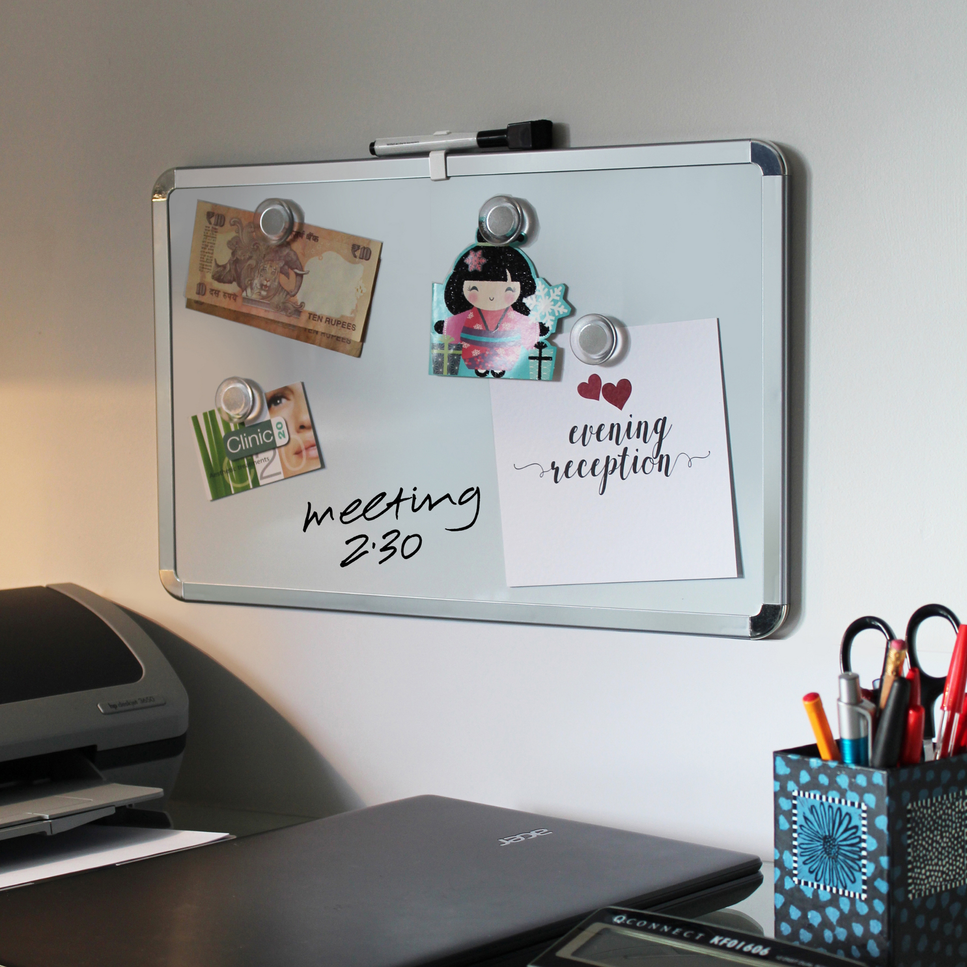 A 28x43cm dry erase board with chrome corners is mounted on a wall, displaying various notes and magnets. Items on the board include a ten rupee note, a cartoon shaped card, a clinic appointment card, wedding invitation, and a handwritten note saying 'meeting 2:30'. A closed laptop and a container holding pens and scissors sit on a desk below the board, displaying the versatile applications of the whiteboard.
