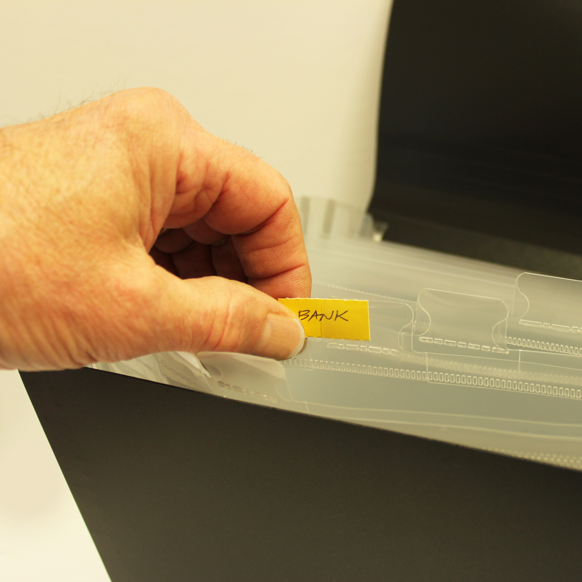A person's hand is inserting a yellow tab labeled 'BANK' into a clear plastic sleeve of an expanding file organizer. The action suggests categorizing or labeling documents for efficient organization, with the hand positioned for precise placement of the label.