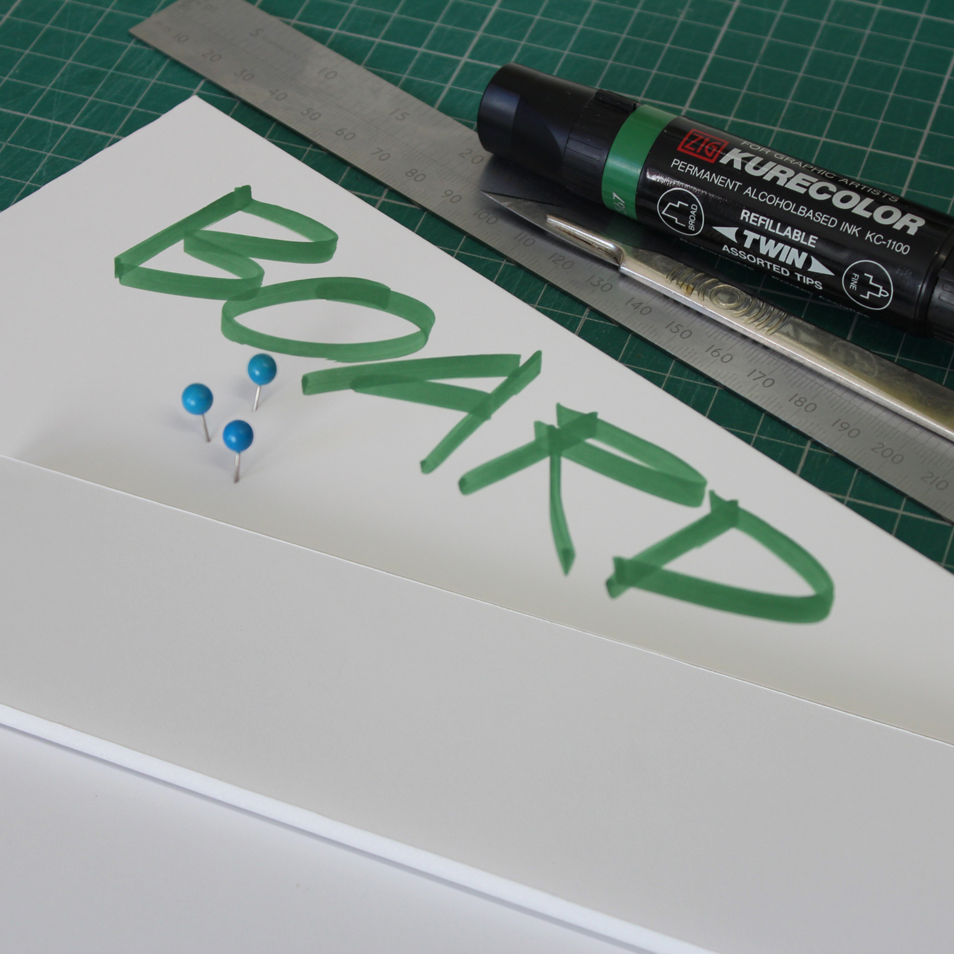 Creative display of white 5mm foam board on a cutting mat. One sheet has been cut into a long strip, and sits on top of a complete sheet, which has 'BOARD' handwritten on it in green marker pen. There are 3 pins stuck into the sheet of foamboard, and a ruler, craft knife and a green marker sit next to the foamboard, displaying the different applications and uses of the product.