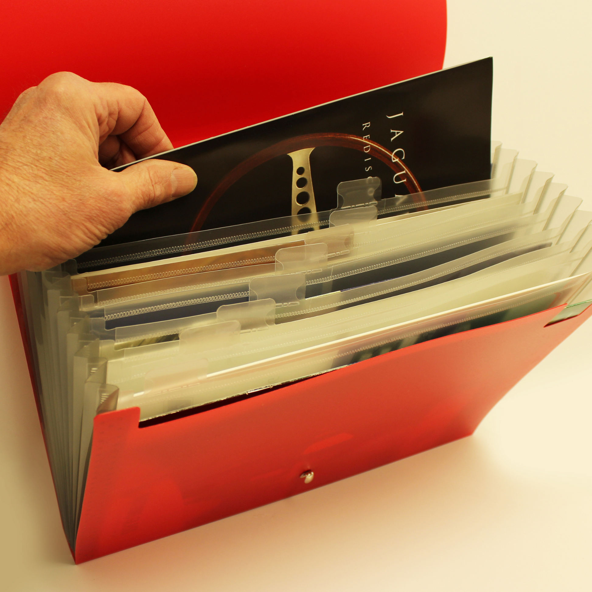 A hand is pulling out a document from a red expanding file organizer with multiple clear dividers and tabs. The visible document has 'JAGUAR' printed on it, suggesting the folder contains professional or automotive-related documents. The organizer is designed for categorization and easy access to contents.