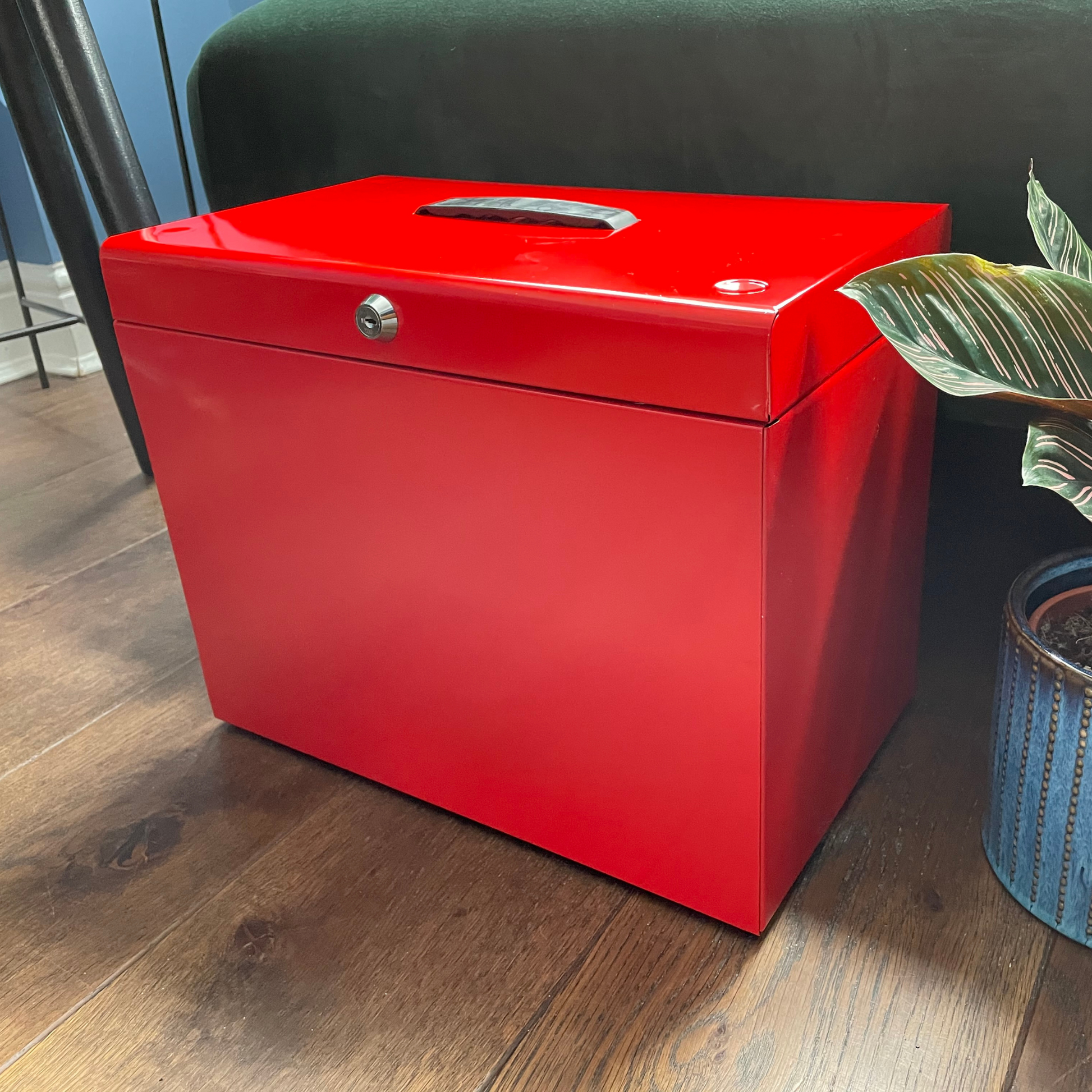 Glossy red A4 steel home file box placed on a wooden floor next to a potted plant, featuring a front lock and a top handle, with five suspension files included for home office organization.