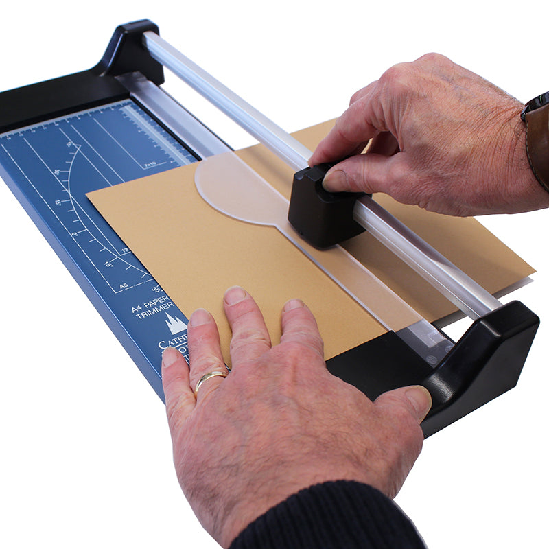 Hands using an A4 rotary paper trimmer to cut a sheet of manila paper, showing the trimmer's blade cartridge and the metal base with measurement guidelines for precise and straight cuts.