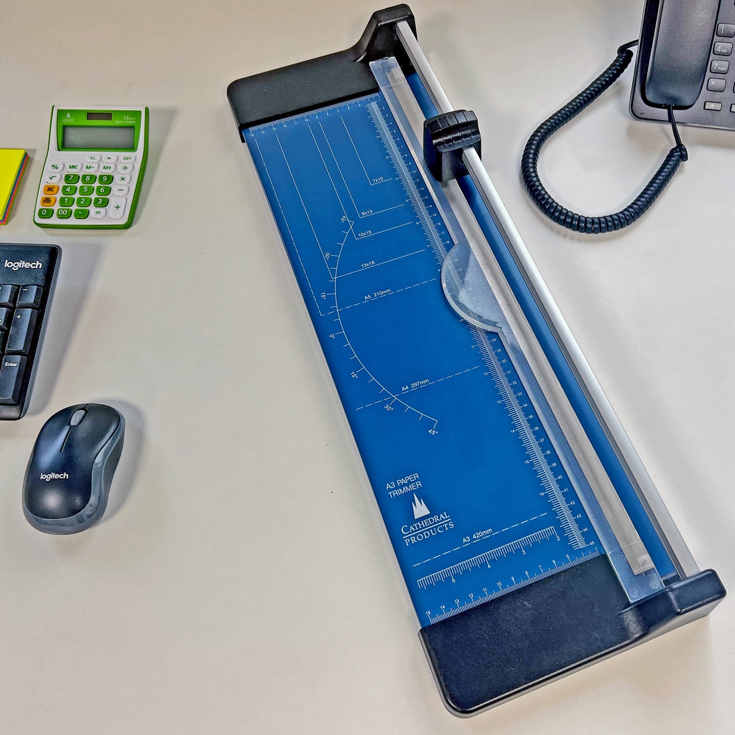 A3 rotary paper trimmer cutter placed on an office desk, featuring a blue metal base with measurement grid, surrounded by office essentials like a calculator, mouse, keyboard, and telephone, illustrating a typical workspace setup.