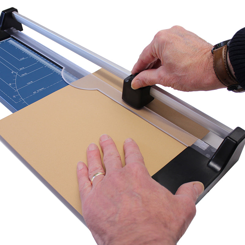 Hands using an A3 rotary paper trimmer to cut a sheet of manila paper, showing the trimmer's blade cartridge and the metal base with measurement guidelines for precise and straight cuts.
