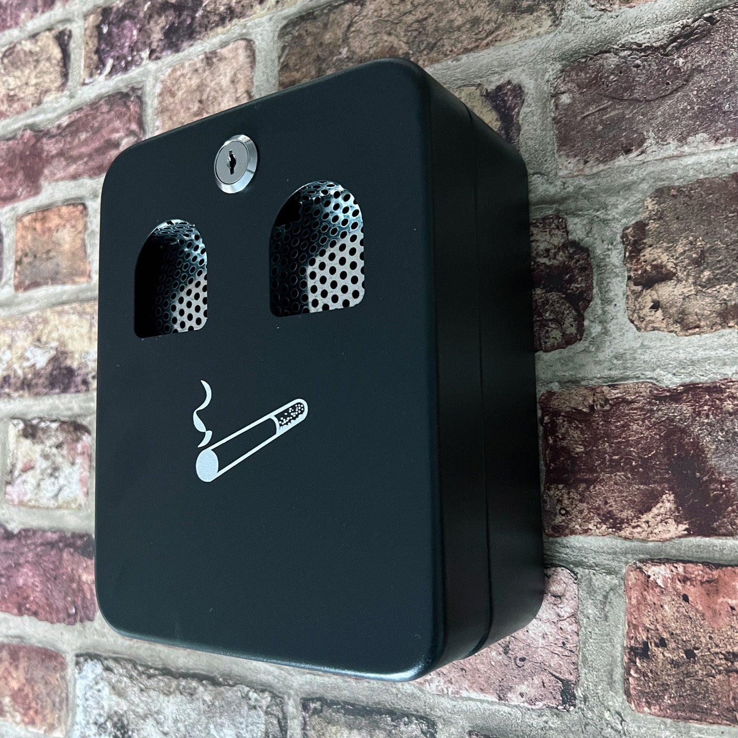 A wall-mounted ash bin with a 1.0L capacity, installed on a brick wall. The matte black bin features a cigarette icon and two perforated metal inserts for cigarette disposal, with a secure lock at the top.