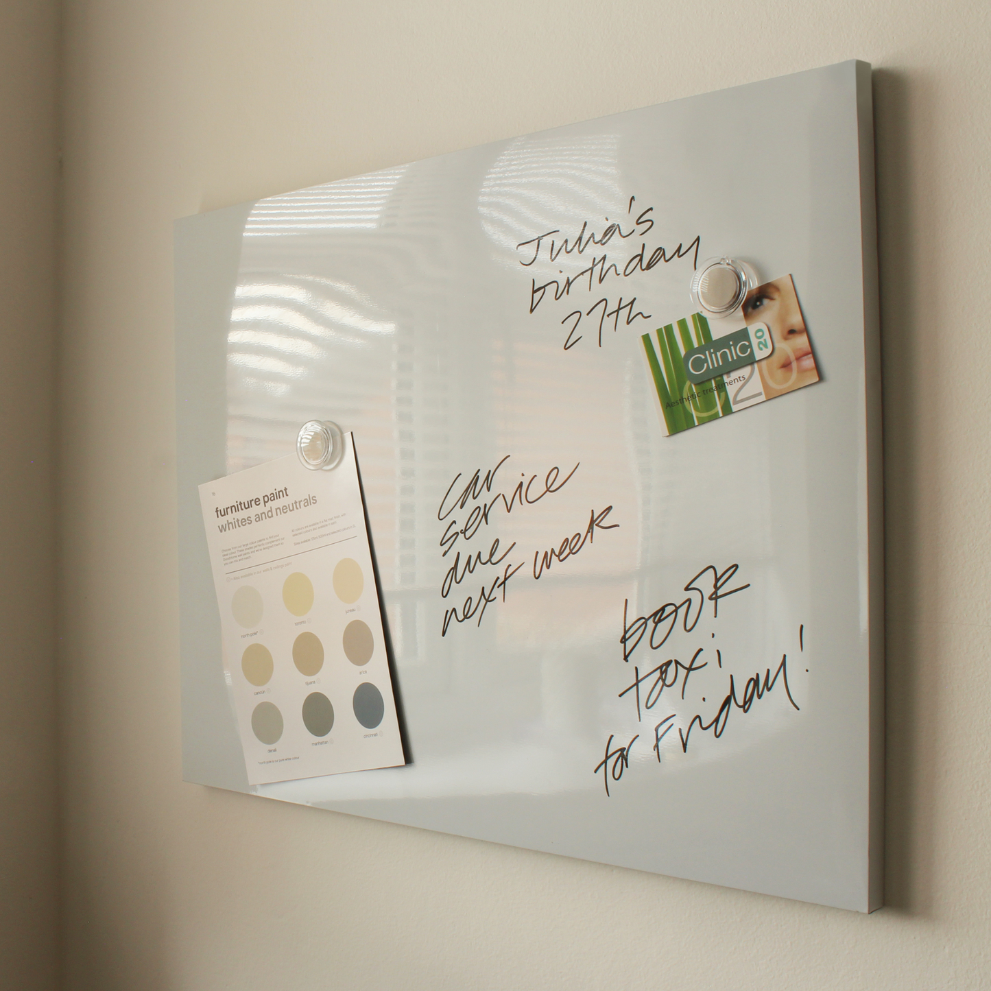 A frameless dry erase board (40 x 51 cm) mounted on a wall with handwritten notes including 'Julia's birthday 27th', 'Car service due next week', and 'Book hotel for Friday!'. Also attached to the board are a furniture paint swatch card and a promotional Clinic flyer.