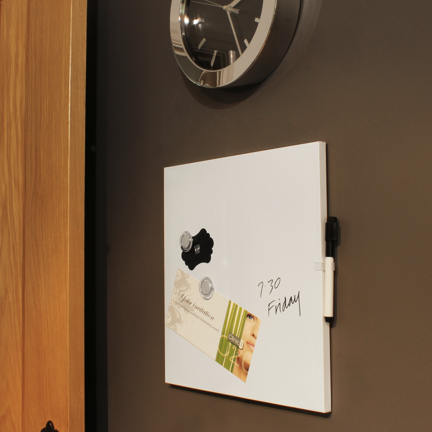 A frameless dry-erase board mounted on a wall with a marker attached, showcasing its use in a modern setting. The board has a wedding invitation and a salon flyer held by magnets, and a handwritten note that reads '7:30 Friday', indicating its role in keeping track of appointments and events.