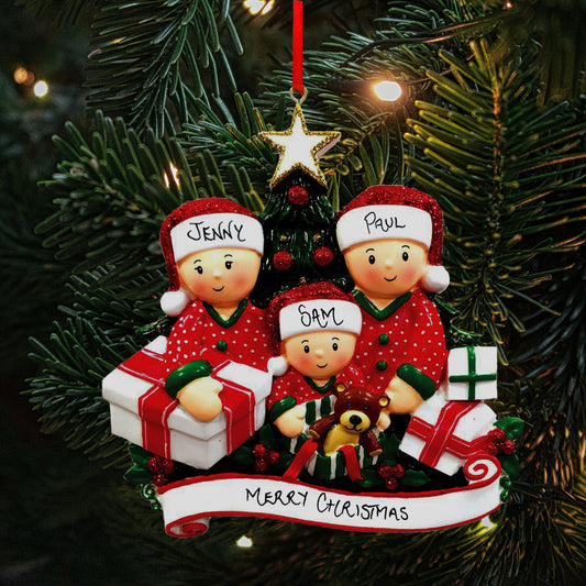 A personalized family Christmas decoration hanging on a tree, featuring a family of three figurines opening presents, labeled 'Jenny', 'Paul', and 'Sam' showing the personalisable capabilities of the ornament. The decoration is adorned with a handwritten 'Merry Christmas' banner and a golden star at the top, set against the backdrop of a Christmas tree's dark green needles.
