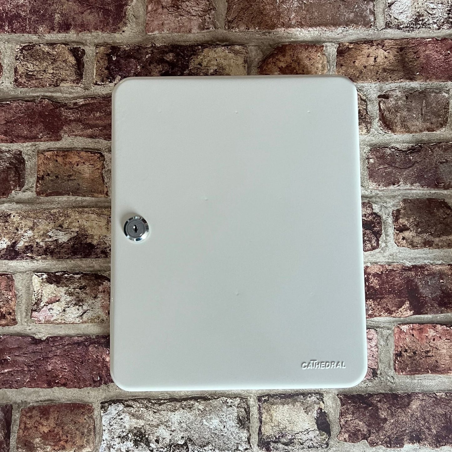 A closed grey Cathedral Products branded metal key cabinet mounted on a rustic brick wall, offering a secure and stylish storage solution for keys.