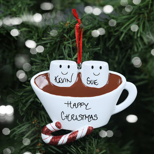 A whimsical personalized couples' Christmas decoration featuring two smiling marshmallow figures in a cup of hot chocolate, labeled 'Kevin' and 'Sue', showing the personalisable capabilities of the ornament. Below them, the cup is adorned with the greeting 'Happy Christmas' and a candy cane, set against the greenery of a Christmas tree with soft light bokeh in the background.