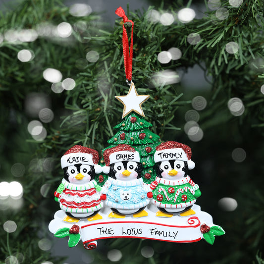 A personalised family of three Christmas decoration featuring three penguins with the names 'Katie', 'James, and 'Tammy' written on their hats, showing the personalisable capabilities of the ornament. They are perched above a banner reading 'Merry Christmas.' The decoration hangs on a Christmas tree, with soft bokeh lights in the background.