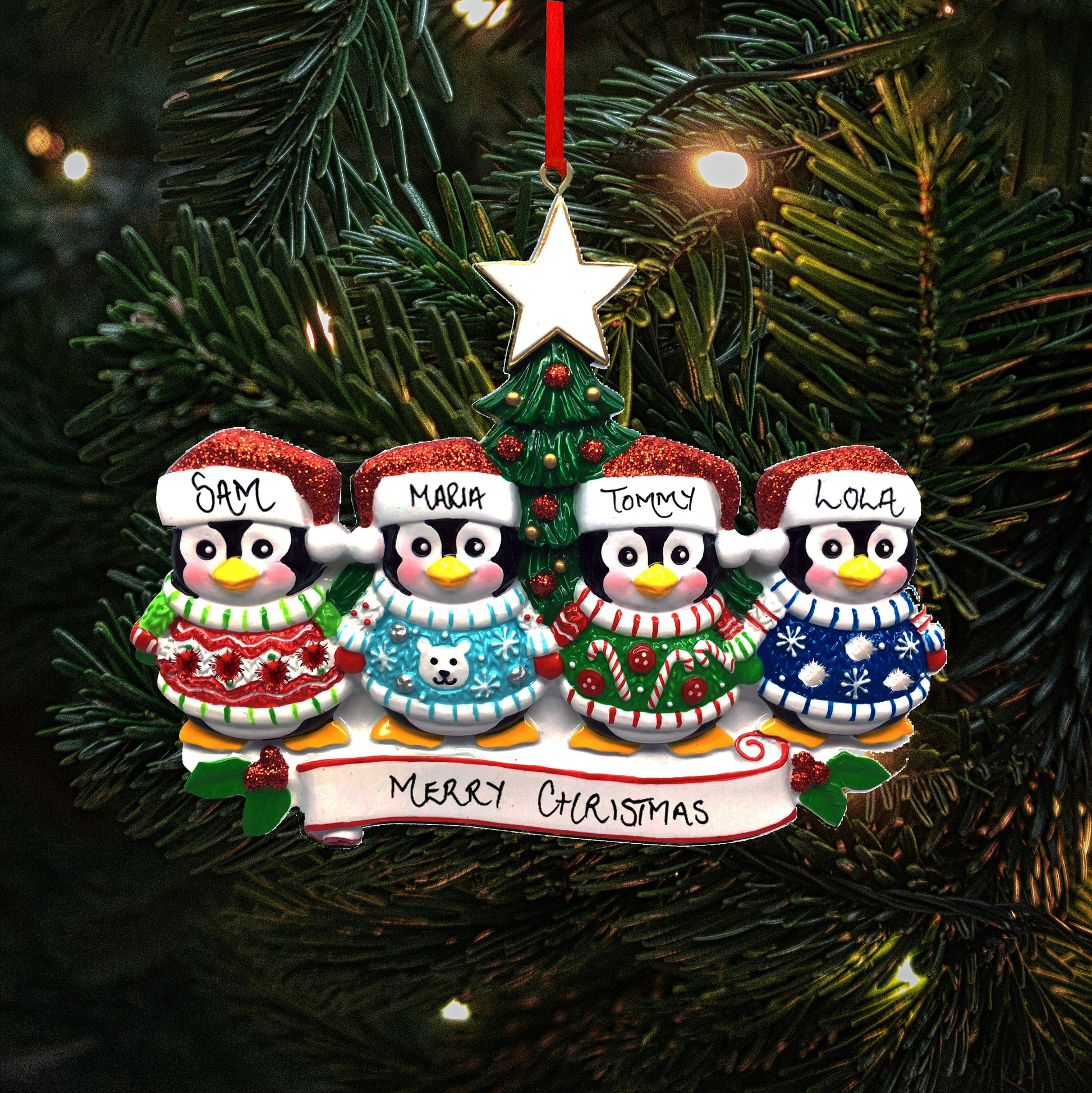A personalized family of four Christmas decoration featuring four penguins with the names 'Sam', 'Maria', 'Tommy' and 'Lola' written on their hats, showing the personalisable capabilities of the ornament. They are perched above a banner reading 'Merry Christmas.' The decoration hangs on a Christmas tree, with lights in the background.