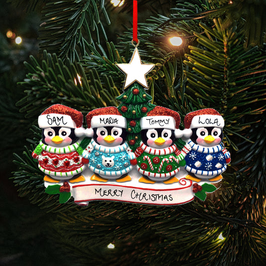A personalized family of four Christmas decoration featuring four penguins with the names 'Sam', 'Maria', 'Tommy' and 'Lola' written on their hats, showing the personalisable capabilities of the ornament. They are perched above a banner reading 'Merry Christmas.' The decoration hangs on a Christmas tree, with lights in the background.
