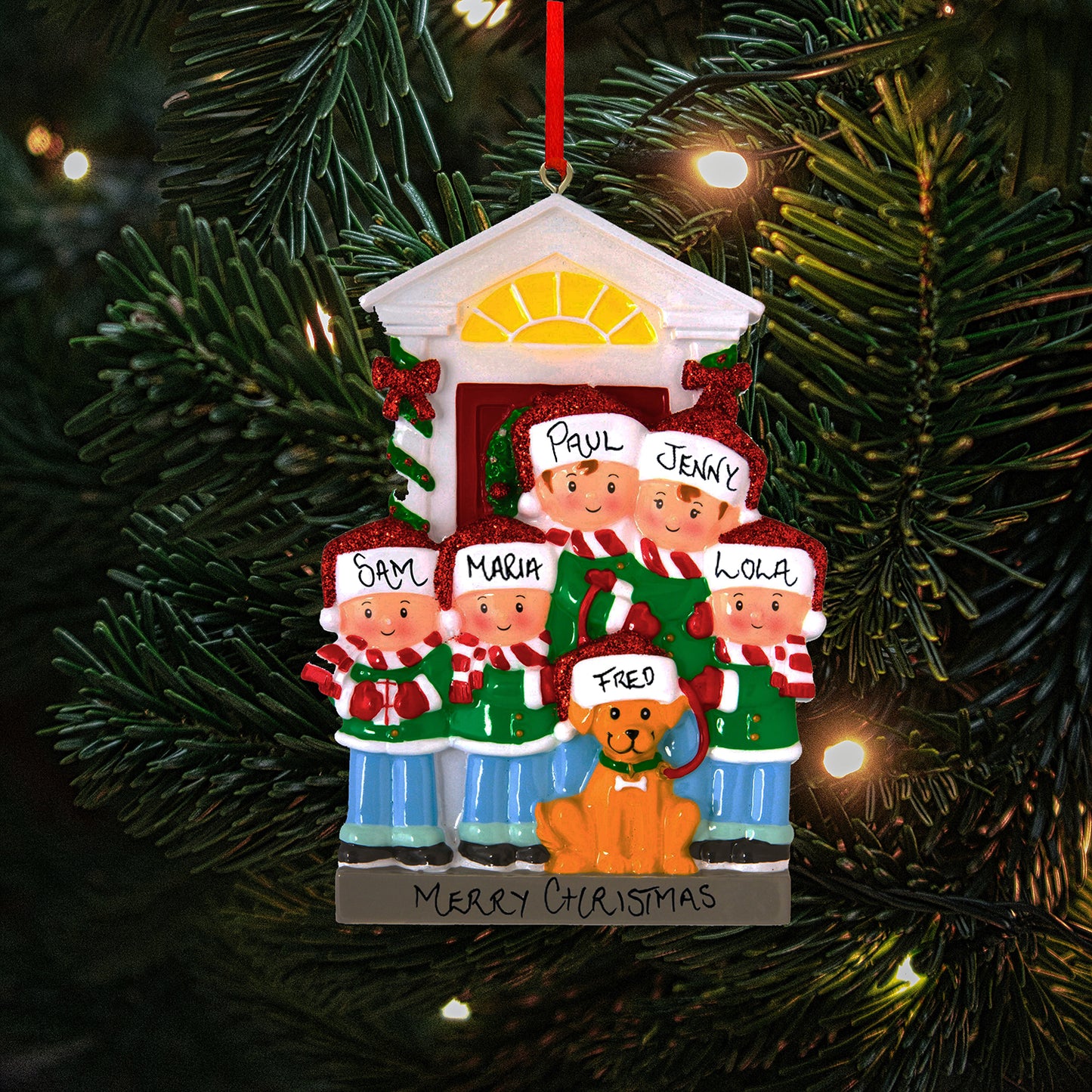 A personalized Christmas tree ornament depicting a family of five with their dog in front of a house, labeled 'Paul', 'Jenny', 'Sam', 'Maria', 'Lola', and 'Fred' on the dog's Santa hat, showing the personalisable capabilities of the ornament. The festive decoration includes a handwritten 'Merry Christmas' banner and is set against a background of a Christmas tree with lights.