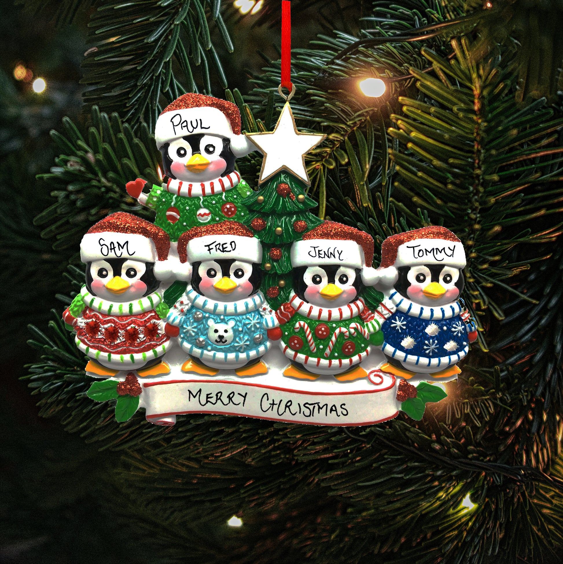 A personalized family of five Christmas decoration featuring five penguins with the names 'Sam', 'Paul', 'Fred', 'Jenny' and 'Tommy' written on their hats, showing the personalisable capabilities of the ornament. They are perched above a banner reading 'Merry Christmas.' The decoration hangs on a Christmas tree, with lights in the background.
