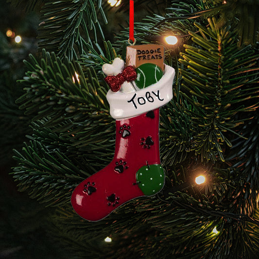 Personalized pet Christmas decoration in the shape of a stocking adorned with a dog bone wrapped in a red glitter ribbon, a green tennis ball and a box of Doggie Treats. The decoration has been personalised with "Toby" in handwritten text on the cuff of the stocking. The ornament is shown against a festive tree background.