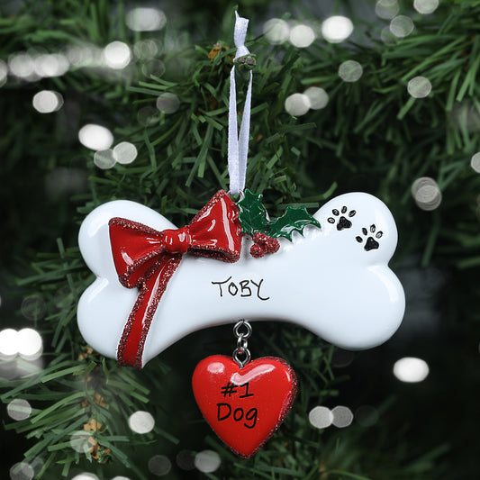 Personalized pet Christmas decoration in the shape of a dog bone adorned with a red bow and a sprig of holly. The ornament also has black pawrints printed on it, and a red heart hanging from the bottom, printed with '#1 Dog". The decoration has been personalised with "Toby" in handwritten text on the bone. The ornament is shown against a festive tree background.