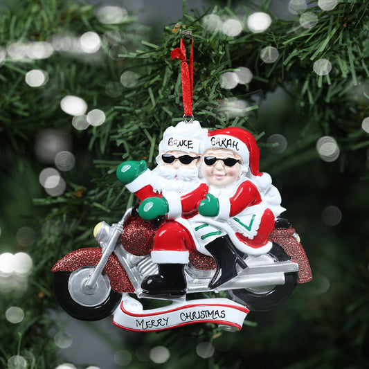 Personalized couples Christmas decoration featuring biker Mr. & Mrs. Claus on a motorcycle, with names 'Bruce' and 'Sarah' written on their hats, and 'Merry Christmas' on the base, showing the personalisation aspect of the decoration, against a festive tree background.