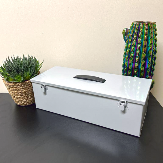 A light grey multi-use hobby and tool box with a double toggle closure and black plastic fold down handle is placed on a dark table next to a potted succulent in a woven basket, and an irridescent cactus vase.