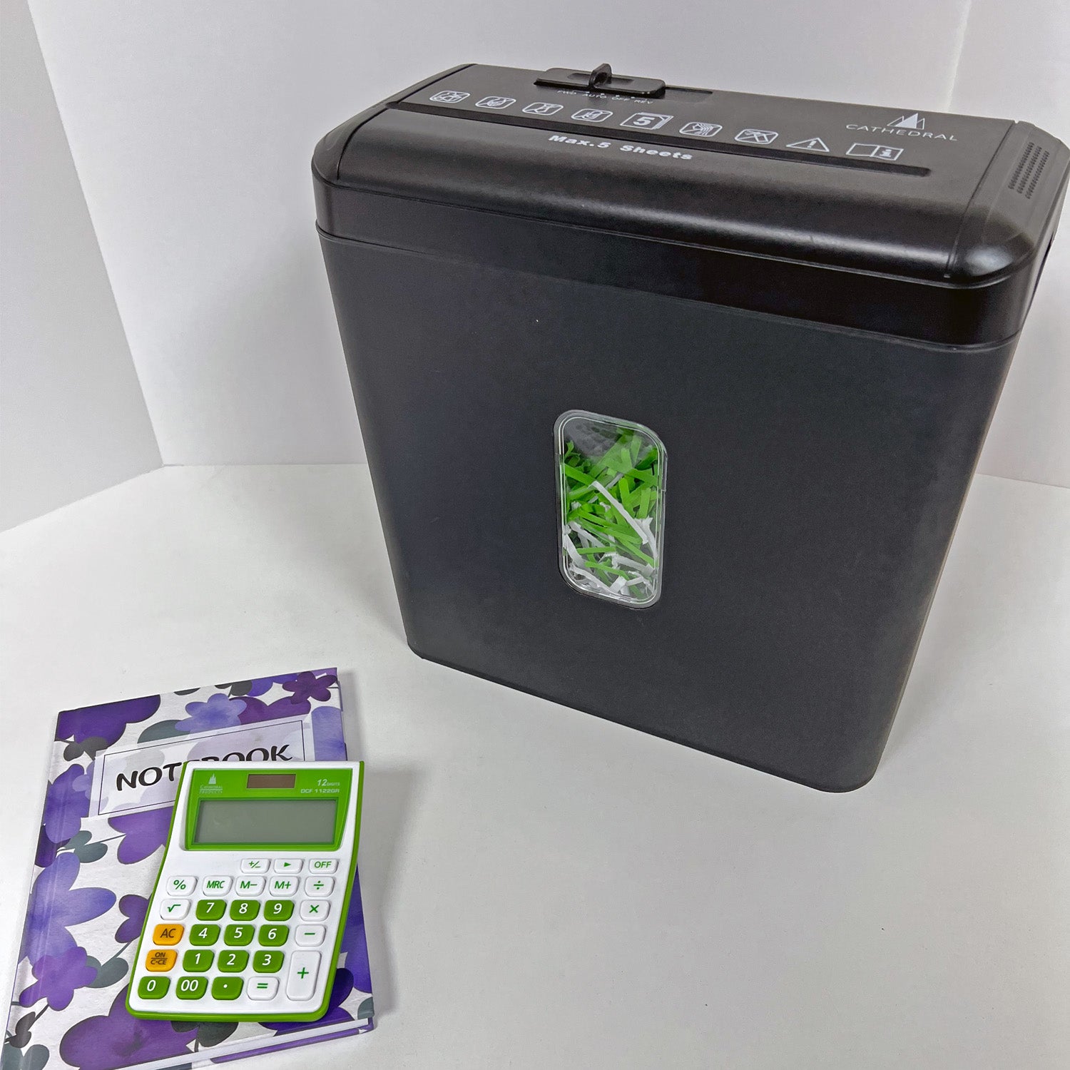 Black Cathedral Products cross cut paper shredder with shredded paper visible in the front window, accompanied by a purple floral notebook and a green calculator, on a white background.