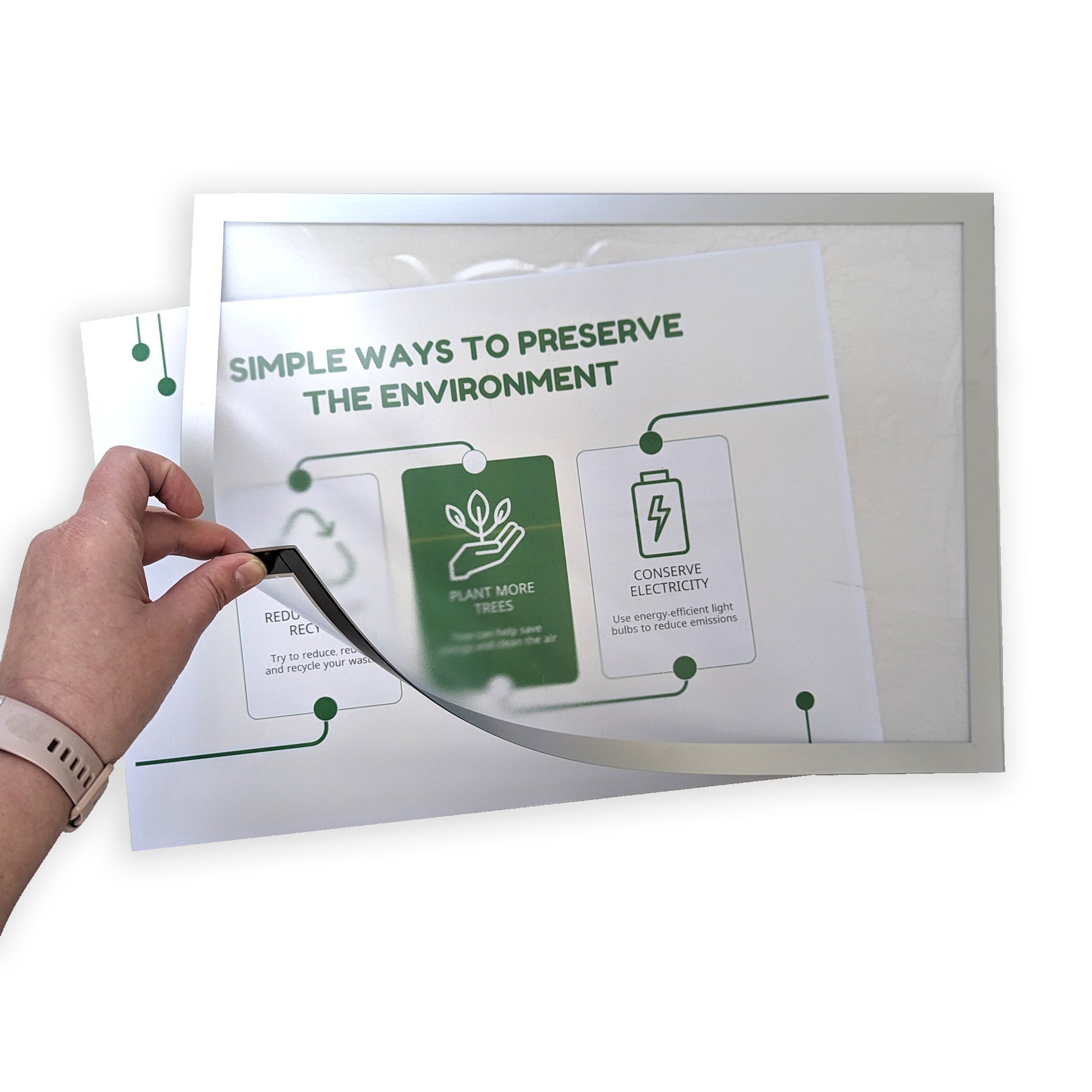 A person's hand is shown peeling back the corner of an A3 self-adhesive magnetic display frame to insert a poster with the message 'SIMPLE WAYS TO PRESERVE THE ENVIRONMENT', featuring eco-friendly tips such as 'PLANT MORE TREES' and 'CONSERVE ELECTRICITY'.